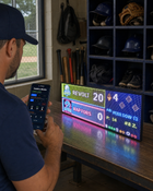 FieldFire Standard three-panel LED matrix scoreboard in a baseball clubhouse with FieldFire Mobile Manual Control on a phone.
