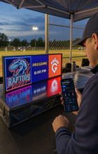 FieldFire Standard three-panel LED matrix scoreboard at a protected tournament desk with FieldFire Mobile Manual Control on a phone.